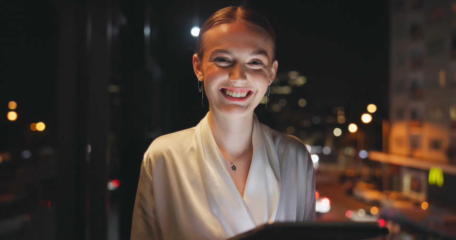 An agent smiling as she checks her phone from a balcony at night, city lights behind her.