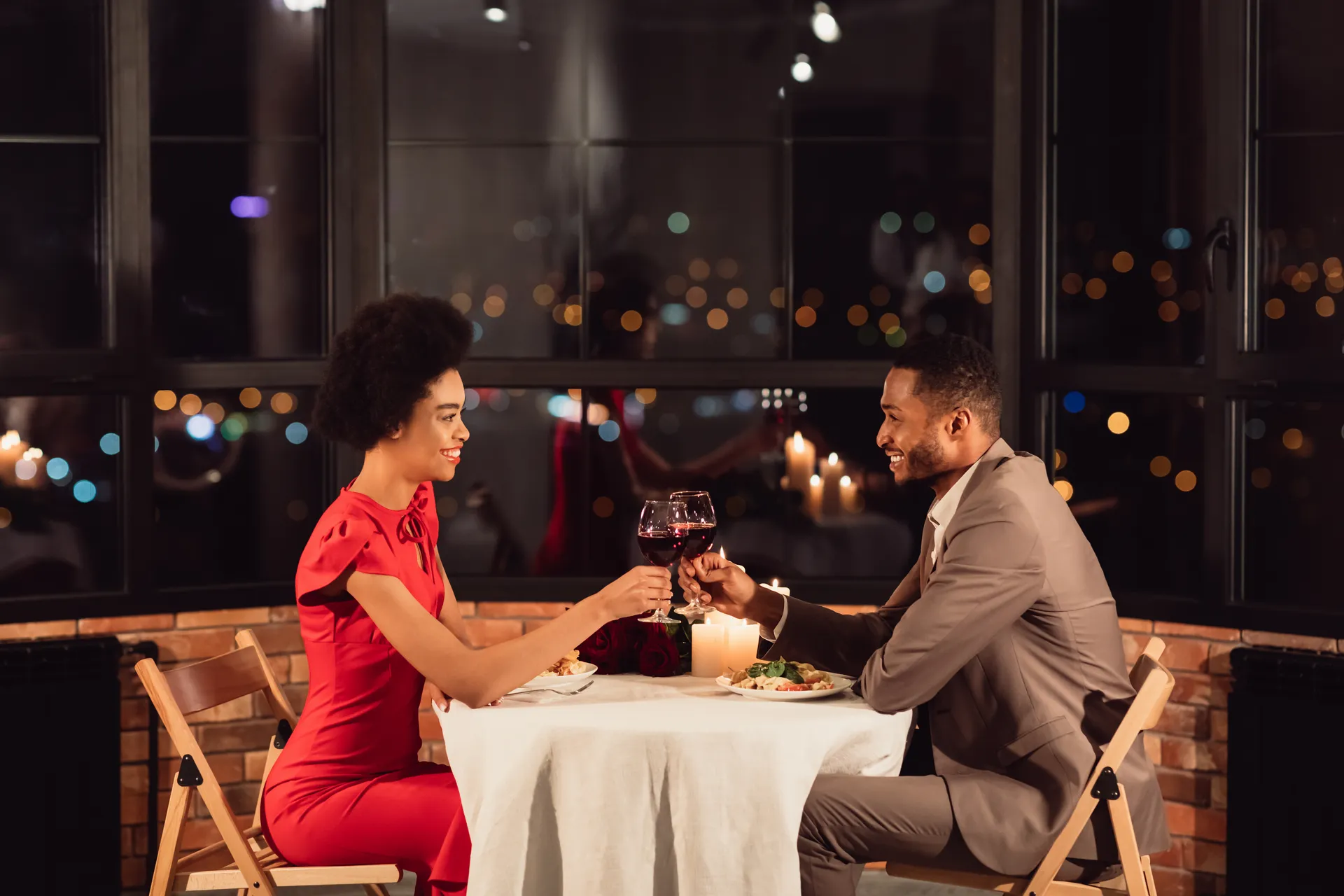 A couple toasting wine at a candlelit dinner table, city lights through the window behind them.