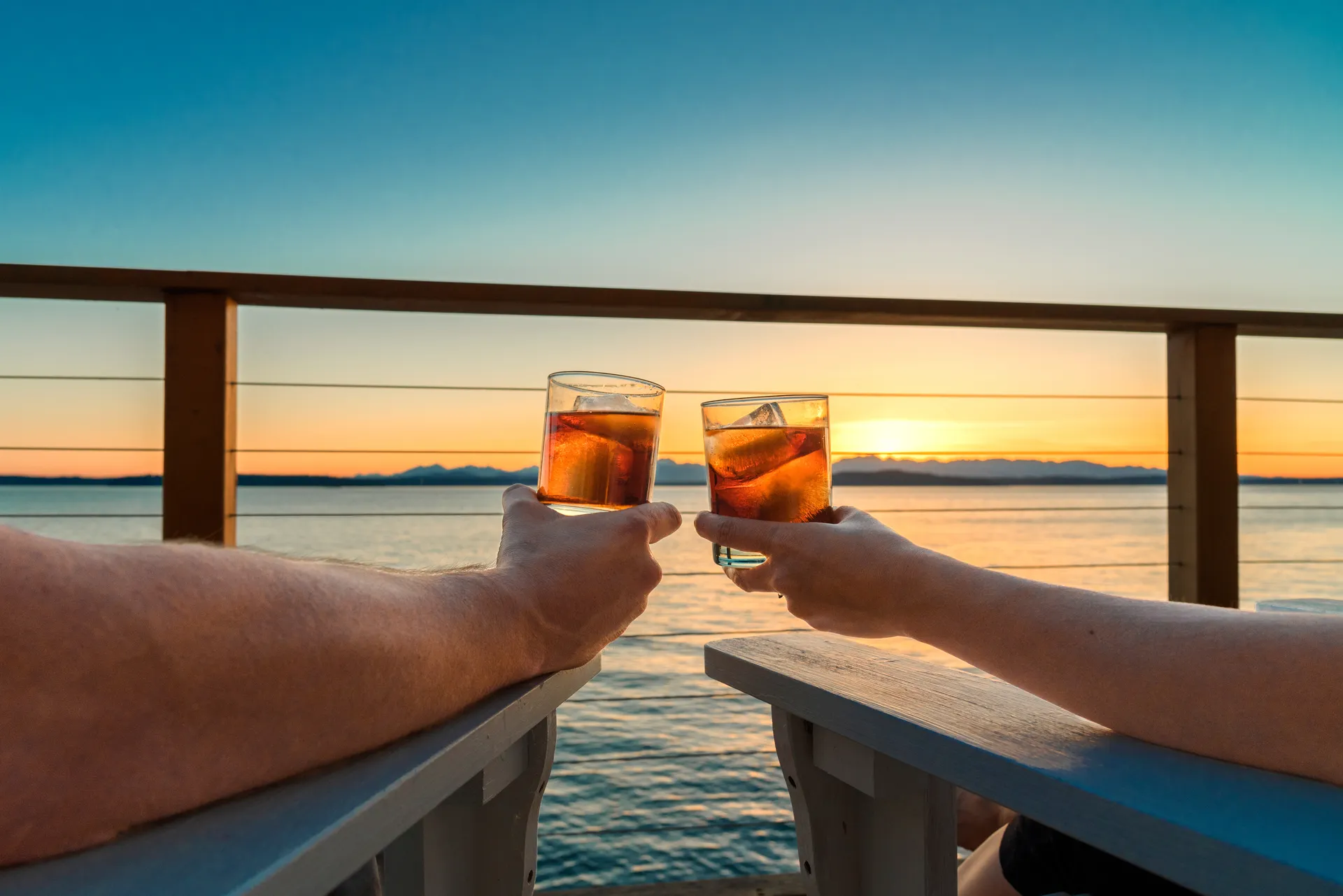 Two hands toasting drinks over a railing at sunset, water and mountains in the distance.