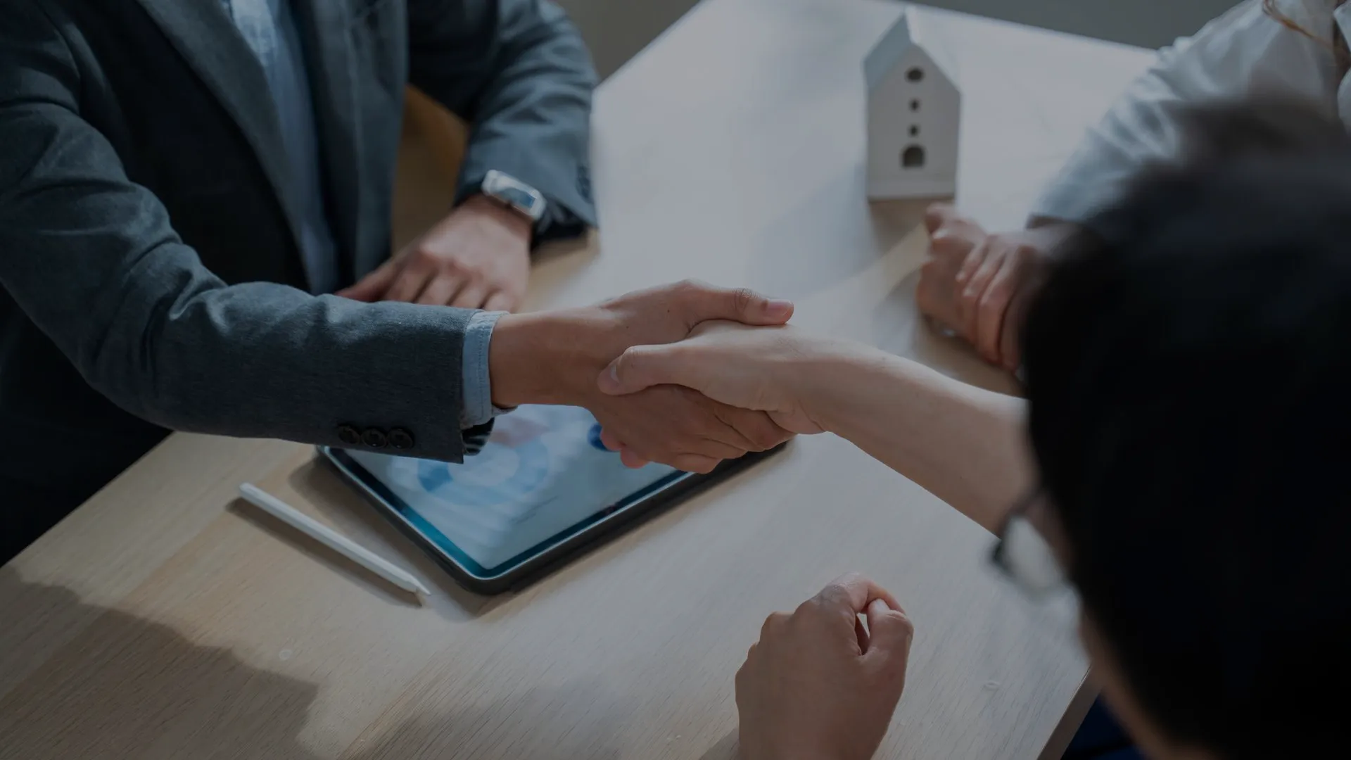 A closing handshake over a desk with a tablet, pen, and a small house model, the paperwork of a deal in progress.