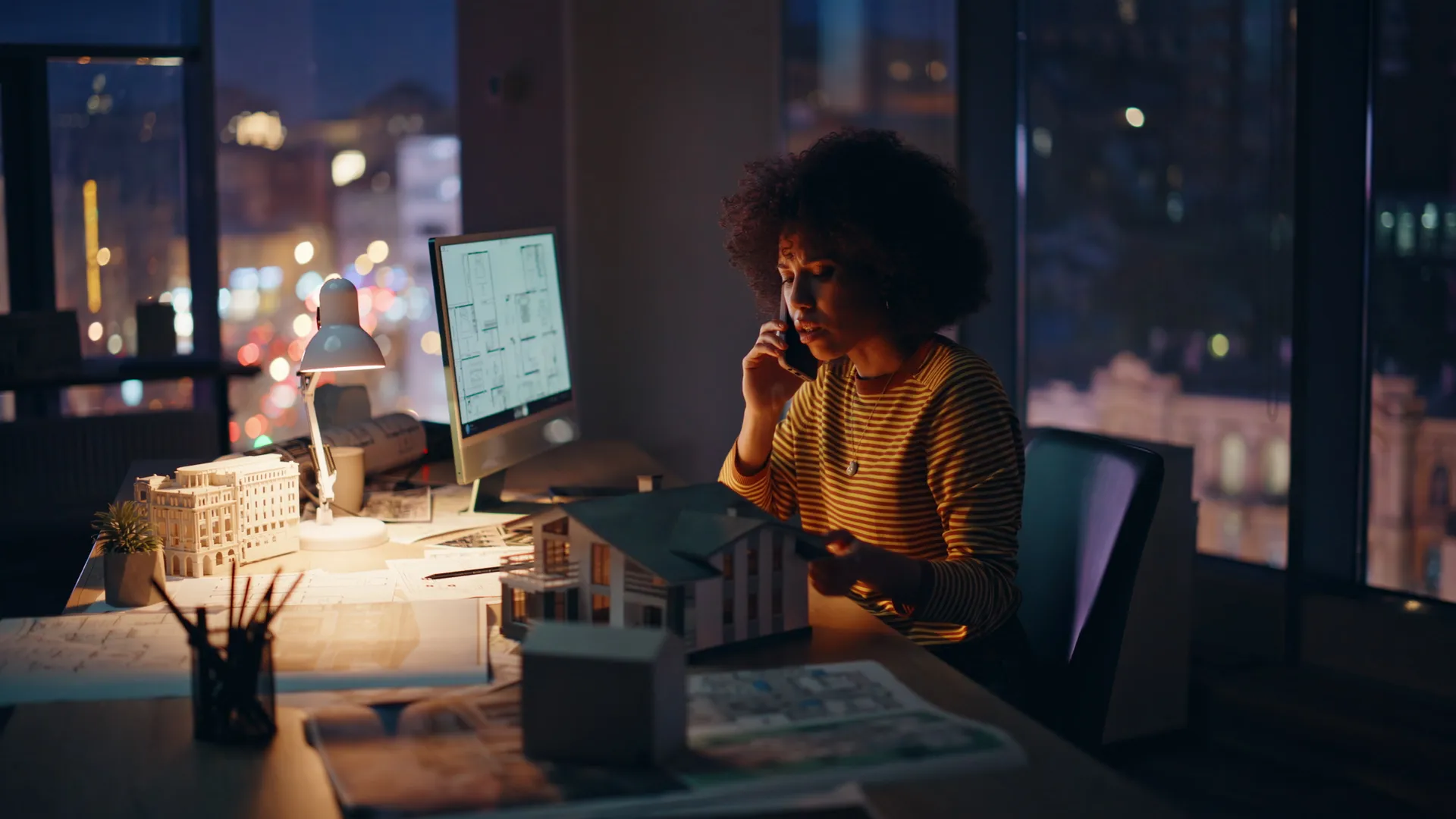An agent on a phone call late in the evening, cool city light through the window and a warm desk lamp lighting her workspace.